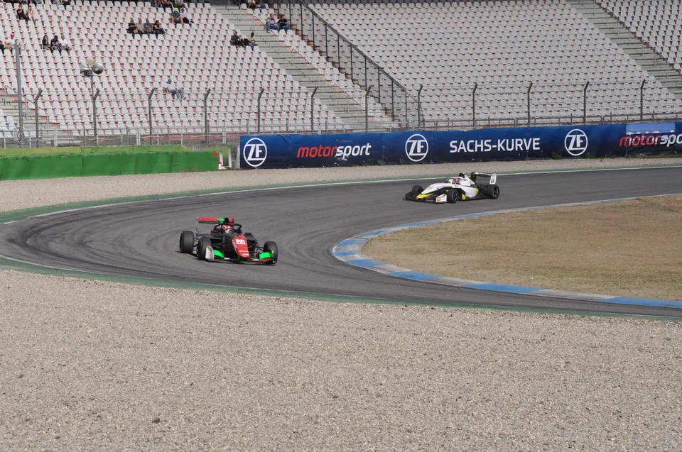 Red and green single-seater ahead of a white and yellow car at the Sachs-Kurve