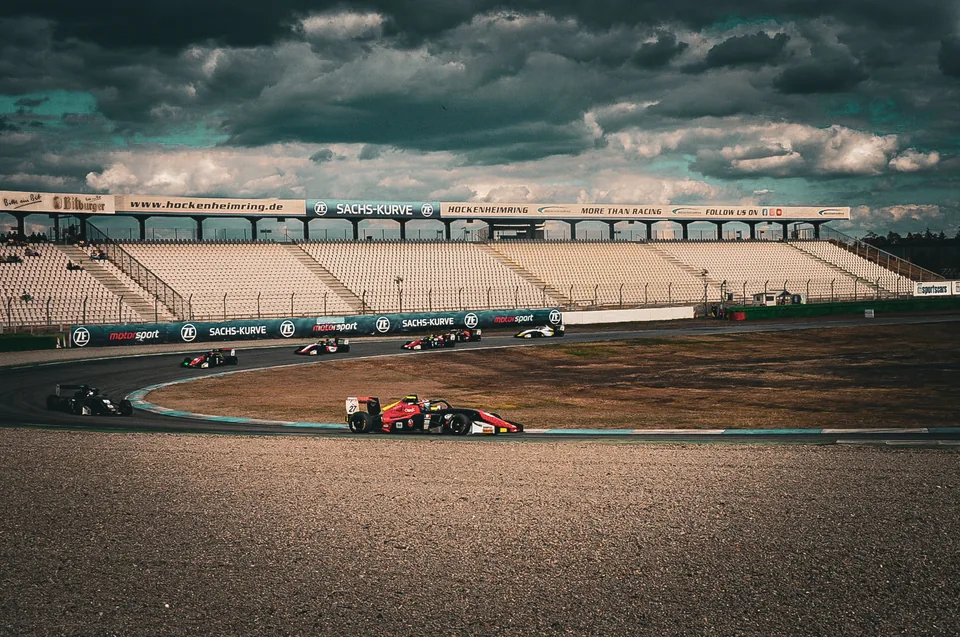 A line of single-seater race cars running through the stadium section under a dark sky