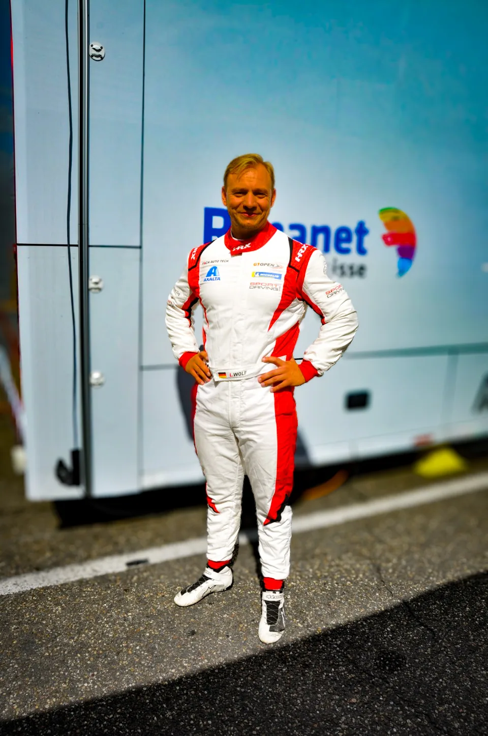 Driver in white and red race overalls standing in front of a team transporter