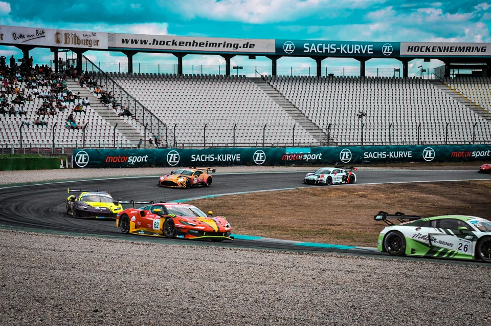 Five GT3 cars running through the Sachs-Kurve with spectators in the grandstand behind