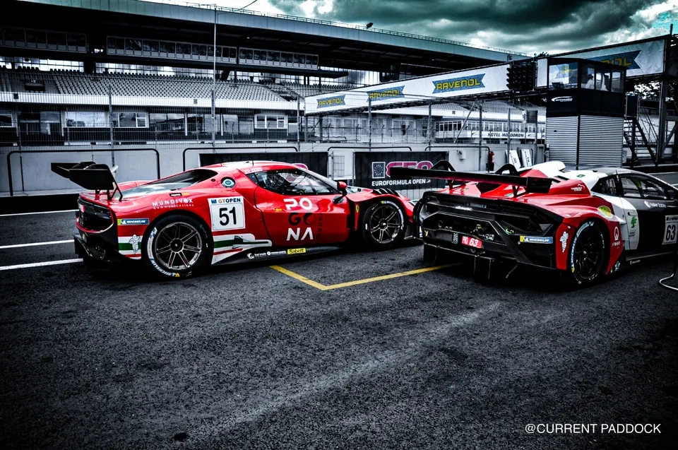 Red number 51 Ferrari and number 63 Lamborghini standing in the pit lane under heavy cloud