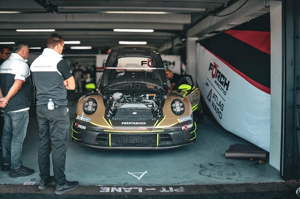 Gold and green Porsche 911 race car in a garage with its bonnet raised and crew standing nearby