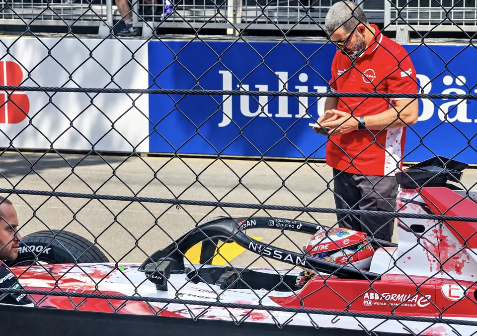 Nissan Formula E car on the grid with a team member checking a phone beside it, seen through catch fencing