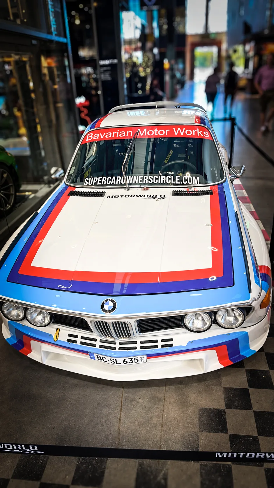Front of a striped BMW racing coupe with a Bavarian Motor Works windscreen banner, shot down the length of the hall