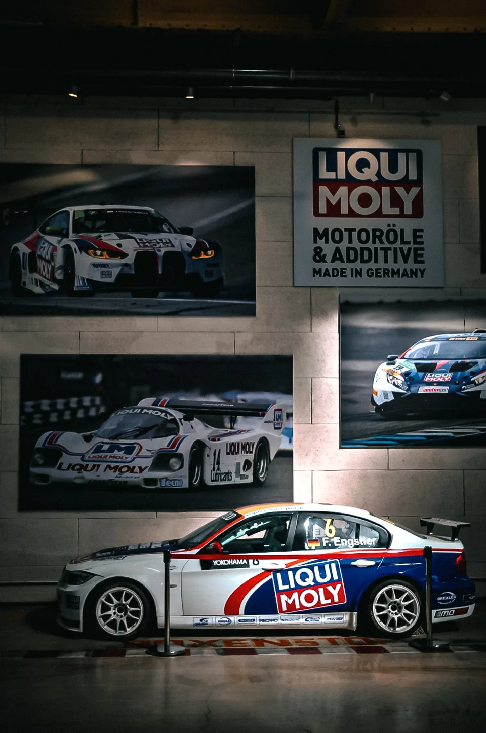 White and blue touring car parked below three large motorsport photographs on a stone wall